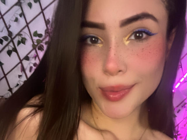 Lara Mancini, met lang bruin haar, blauwe eyeliner, gouden oogschaduw, roze lippen, sproeten en een subtiele glimlach, toont haar gezicht en bovenborst. Borsten zijn gedeeltelijk zichtbaar..
