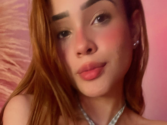Lara Mancini tem cabelo ruivo, lábios rosados e um rosto bonito com um leve sorriso. Ela está usando um colar de prata e tem um pequeno brinco de argola no ouvido esquerdo..