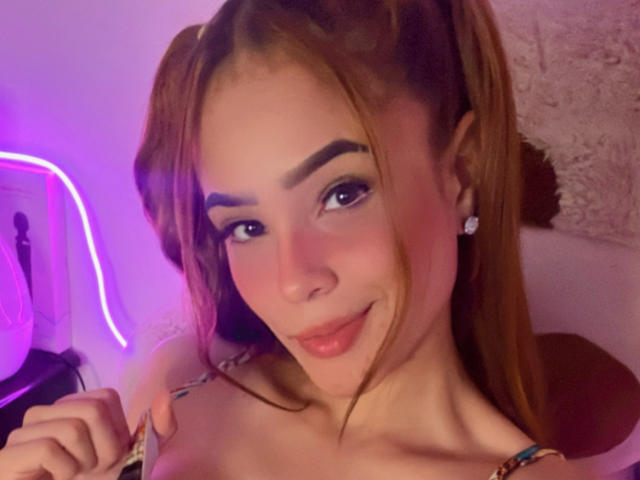 Lara Mancini, con i capelli castano chiaro raccolti in due code, indossa una maglietta colorata, ha l'ombretto azzurro, il rossetto rosa ed è sorridente con una mano vicino al seno. Illuminazione viola tenue..