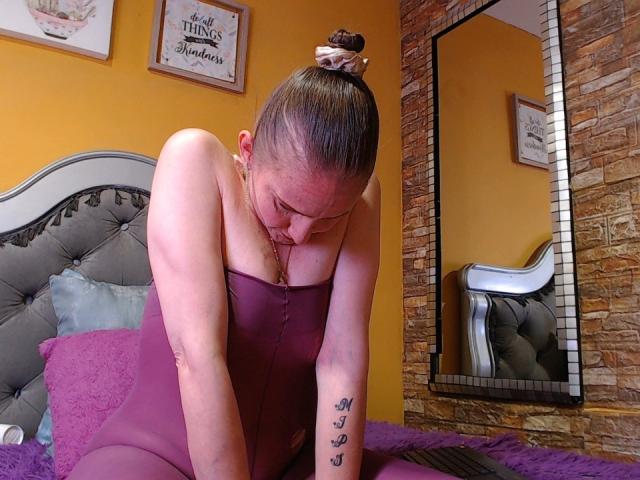 Rubi Anderson, met haar borsten die uit een paarse jurk piepen, armen naar beneden, haar in een knot, tatoeage op haar rechterarm, voorovergebogen, reflectie in een spiegel, stenen muur, oranje muur..