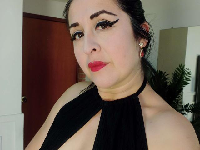ScarletMilfX with dark hair, red lipstick, black halter dress, thick black eyeliner, red earrings, slightly chubby face, and pale skin, standing near a wooden door.