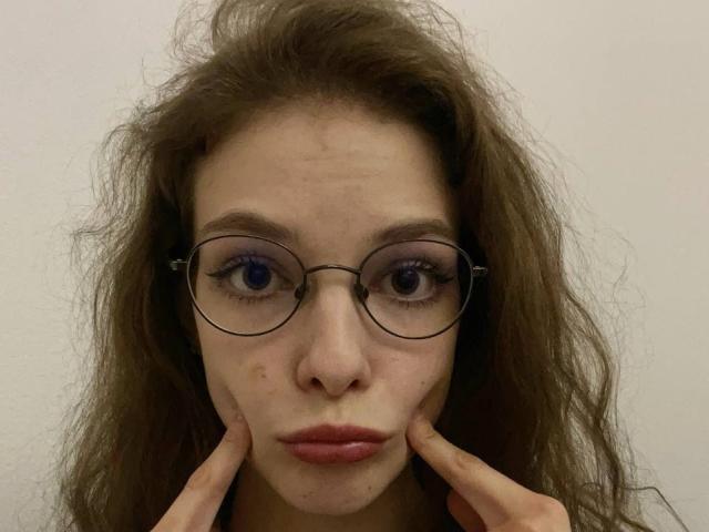 FaithRegulus has messy brown hair, round glasses, purple eyeshadow, and is pouting with fingers on cheeks. She has blue eyes, light skin, and slightly parted pink lips.