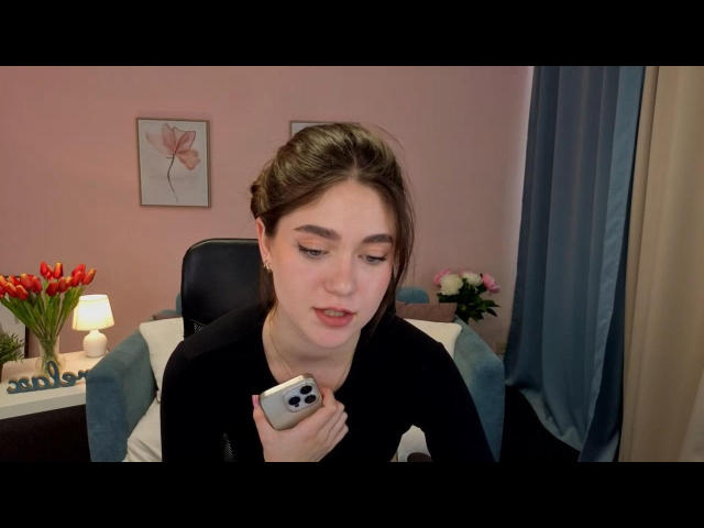 SunnyBloss with brown hair in a black top, holding a silver camera phone, looking down, mouth slightly open, in a pink-walled room with a blue chair and flowers.