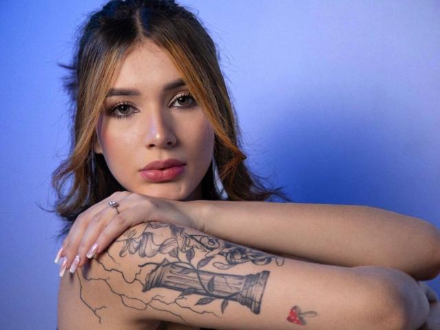 Greta Harris heeft tatoeages op haar rechterarm, waaronder een vat en een bloemendesign, en een klein rood hart op haar linkerarm. Haar bruine haar is golvend, en ze draagt geen bovenkleding..