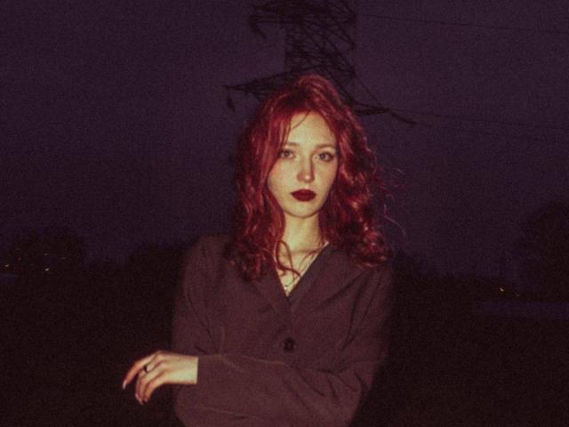 WilliemaeKielar a les cheveux roux et bouclés, porte un rouge à lèvres foncé et est vêtue d'un blazer noir, les bras croisés, debout devant une silhouette sombre de lignes électriques la nuit..