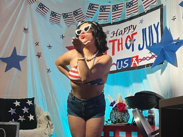 RoxanneHan with curly black hair, white heart sunglasses, red lipstick, American flag bikini top, denim shorts, hands near face, standing, blue lighting, star and flag bunting, black hat.