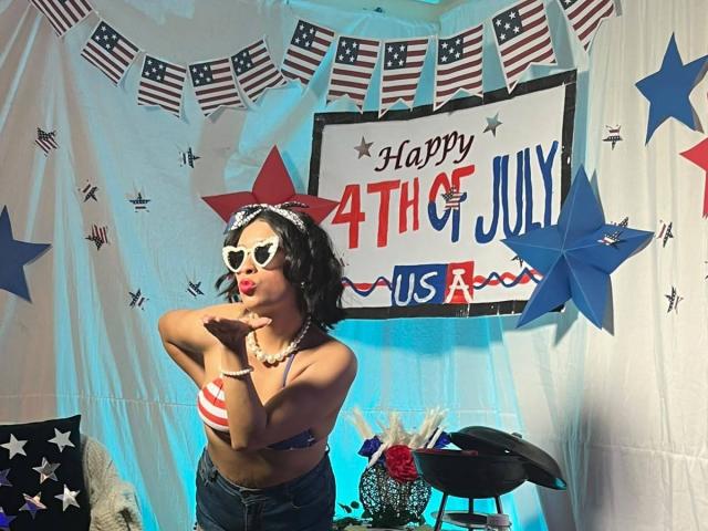 RoxanneHan with black curly hair, wearing white heart-shaped sunglasses, red star headband, striped bikini top showing small boobs, blue jeans, and pink lipstick, posing with hands near face.