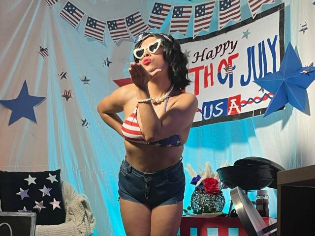 RoxanneHan with tits in a striped bikini top, wearing denim shorts, white star sunglasses, and a pearl necklace, posing with hands near mouth, in a patriotic-themed setting.