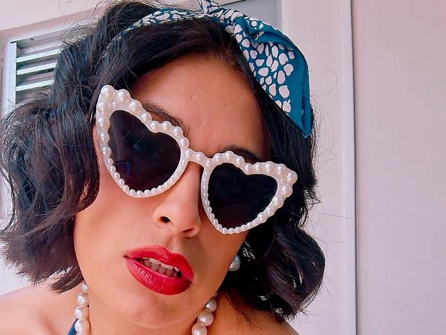 RoxanneHan with curly black hair, wearing heart-shaped sunglasses with pearl trim, blue floral headscarf, red lipstick, and pearl necklace, looking seductive with slightly parted red lips.