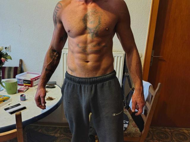 LuciferSun es un hombre musculoso, sin camisa y con tatuajes, que lleva pantalones de chándal grises y está de pie en una habitación con una mesa y una silla. Se pueden ver sus abdominales y su pecho..