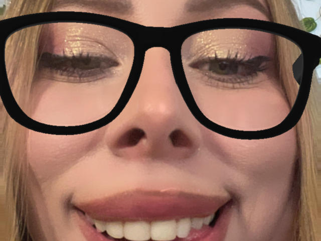 Il viso di JennyLawry, con occhiali neri, ombretto viola brillante e un ampio sorriso che mostra denti bianchi. Le sue labbra rosa sono leggermente dischiuse. I capelli biondi incorniciano il suo viso..