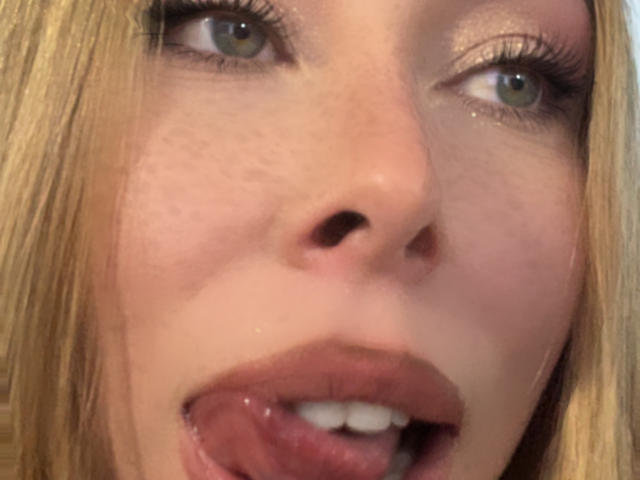 Jennys Gesicht, blondes Haar, grüne Augen und leicht geschwollene, rosafarbene Lippen, wobei ihre Zunge herausragt. Ihre Nase ist mittig platziert, und ihr Make-up ist dezent..