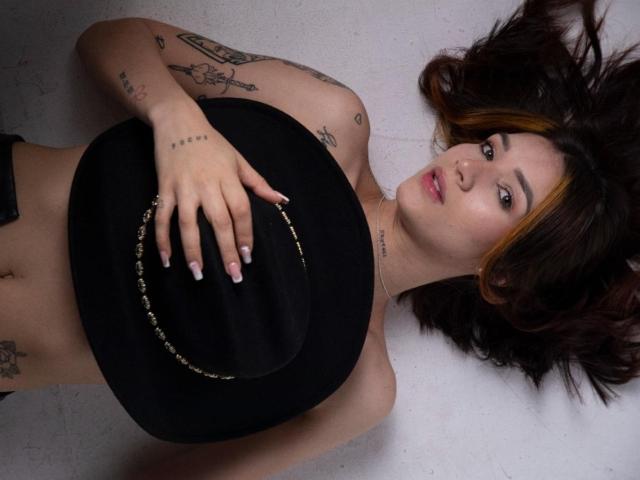 GretaHarris lying on her back, holding a black hat with a gold chain, tattoos on her arms, dark brown hair with blonde highlights, looking at the camera.