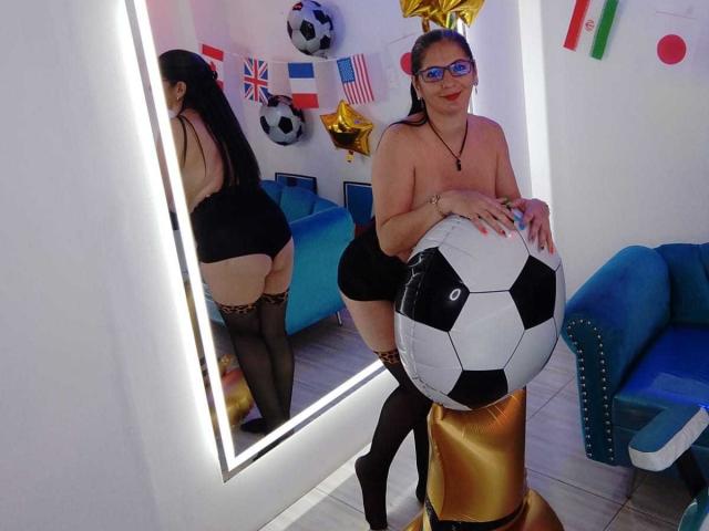 MadameScarlett with long black hair, glasses, and red lipstick, topless, wearing black panties and thigh-high stockings, leaning on a soccer ball in a room with a mirror.