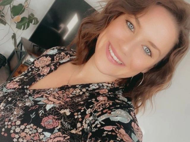 HeyLina-hot with wavy brown hair, blue eyes, and a smile, wearing a floral black shirt, showing her tits subtly in a selfie. She's got a cute, sexy look.