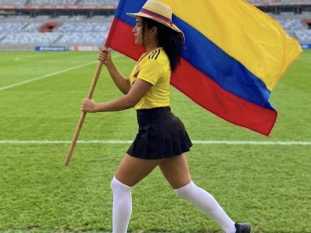 SophieSonyas, avec ses cheveux noirs bouclés, sa chemise jaune, sa jupe noire, ses chaussettes blanches, son chapeau de paille, tenant le drapeau colombien, marchant sur un champ vert, montrant ses fesses en vue de profil..