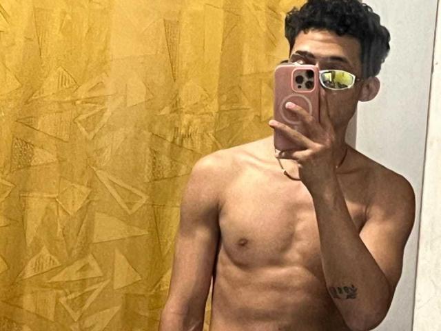JaredHt está sem camisa, segurando um telefone rosa, com uma tatuagem no braço direito, em frente a uma cortina amarela e texturizada, mostrando um corpo superior tonificado e esguio..