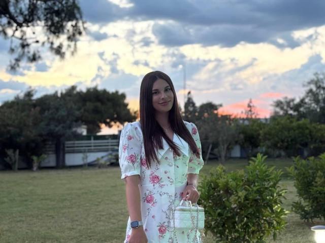 BlackFatal, with long brown hair, wearing a white floral dress, holding a glass, stands in a grassy yard, sunset sky, dark clouds, and trees in the background.