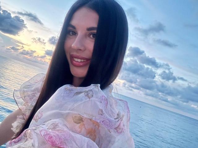 BlackFatal with long brown hair, pink floral sheer top, smiling, showing her teeth, standing by the ocean at sunset, blue sky with clouds, gentle water, cute, sexy.