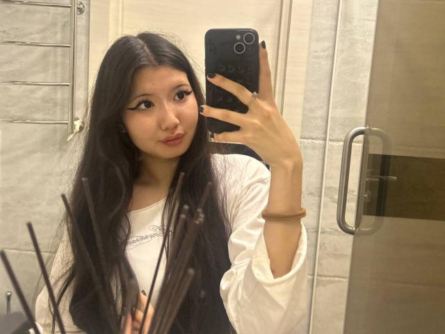 BernadineFray, avec de longs cheveux noirs, portant une chemise blanche et un gilet noir, prend un selfie dans une salle de bain, tenant un téléphone noir, montrant des ongles noirs et un bracelet marron..
