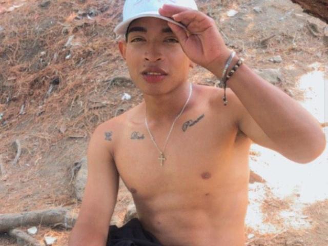 Shirtless ChrisSexi with tan skin, tattoos on chest, wearing white cap, cross necklace, and bracelets, slightly open mouth, right hand touching cap, sitting on dirt, pine needles.