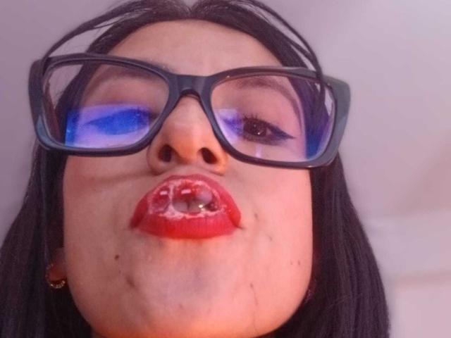 ZarayFeet mit schwarzen Brillen, blauem Augen-Make-up und rotem Lippenstift hat eine Perle im Mund, blickt nach unten mit leicht geöffneten Lippen und hat schwarzes Haar..
