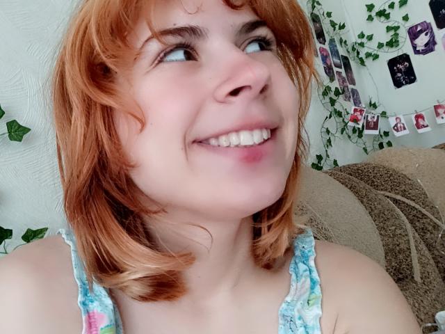 MireilleSpotwood with red hair, smiling up, showing teeth, wearing a floral tank top, looking sideways, fair skin, cute, small nose, blue eyes, medium-sized breasts, casual selfie.