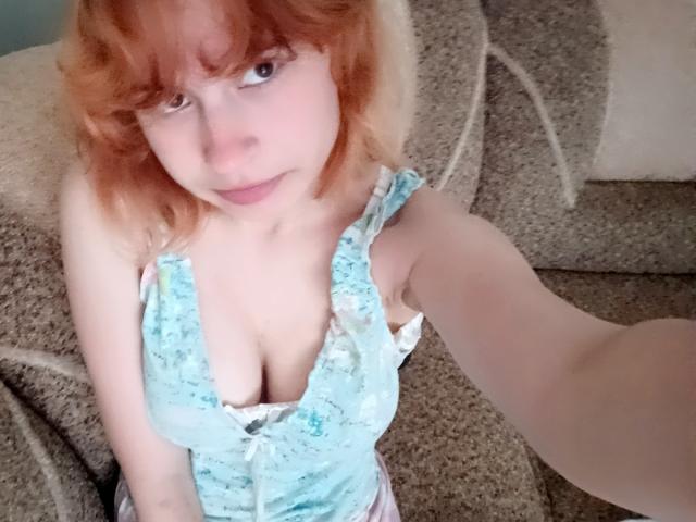 MireilleSpotwood with red curly hair, pale skin, and blue floral top, showing her cleavage in a selfie, arms slightly raised, on a beige, textured couch.