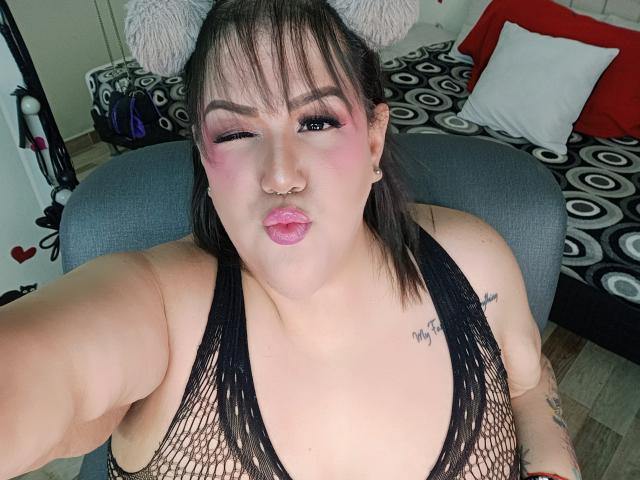 GiiannaMorrys, a curvy woman with pink makeup, nose ring, and tattoos, puffs her pink lips while wearing a black fishnet top, showing her tits. She's winking.