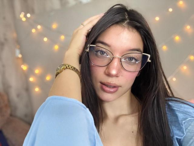 MichellFord met een bril, lang donker haar, lichte huid, gekleed in een blauw shirt, een gouden horloge, die haar haar aanraakt, roze lippen, subtiele make-up, zacht licht, lichtjes in de achtergrond..
