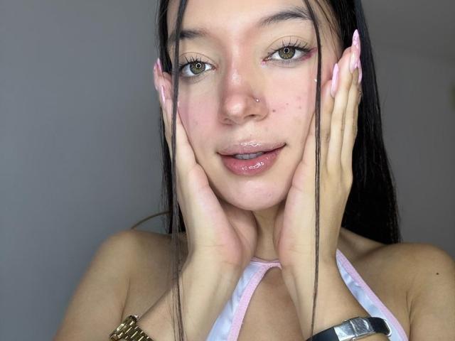 MichellFord, com lábios rosa brilhantes, piercing no nariz e cabelos longos e escuros, mãos nas bochechas, olhos verdes, unhas rosa, vestindo um top rosa de alças, mostrando os seios..