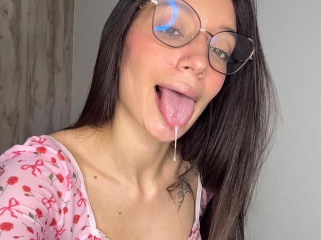 MichellFord with long dark hair, glasses, and pink floral top, tongue out, drooling, showing a small tattoo on her right shoulder, and a nose ring.