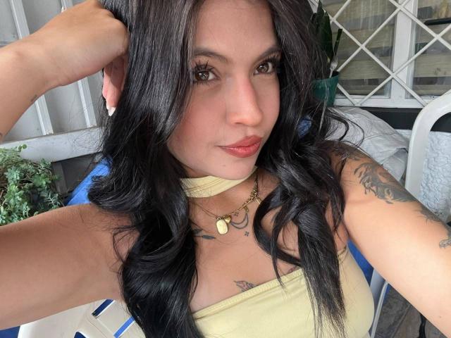 NyraVanel with long black hair, tan skin, and tattoos, wearing a yellow top, looking at the camera with a slight smile, hand in hair, showing off her pouty ass.