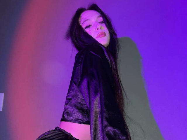 ChaneCarco, con cabello largo y negro, vestida con una blusa de terciopelo negro, mirando hacia atrás con la boca ligeramente abierta, con una luz púrpura y rosa sobre ella. Se puede ver su trasero con unos pantalones oscuros..