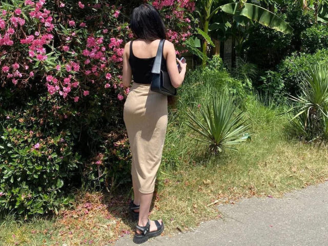 ChaneCarco steht mit dem Rücken zur Kamera, trägt ein schwarzes Oberteil und einen beigen Rock, hält ein Telefon, schwarze Sandalen und eine schwarze Tasche, und befindet sich in der Nähe von rosa Blumen und Grünpflanzen..