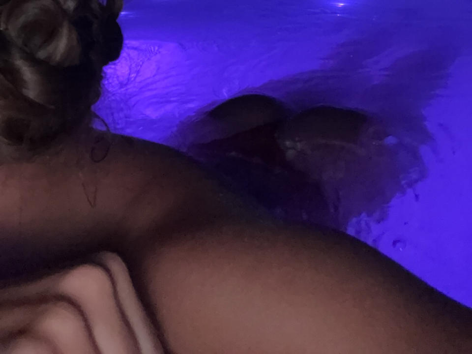 ShashaSxy's bare shoulder and upper chest with a braid, submerged in purple-lit water, showing a partially visible breast and arm. Dark, wet skin and a towel.