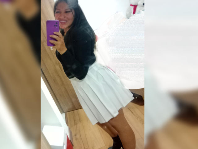 Novarose with long black hair, smiling, wearing a black top and white pleated skirt, taking a mirror selfie with a purple phone, showing her cute ass.