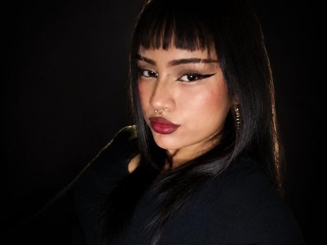 AkariChise ha i capelli neri e lisci, con frangia dritta, un trucco occhi audace con eyeliner ad ali di farfalla, rossetto rosso, un anello al naso e orecchini a cerchio dorati, indossa una maglietta nera, su uno sfondo scuro..