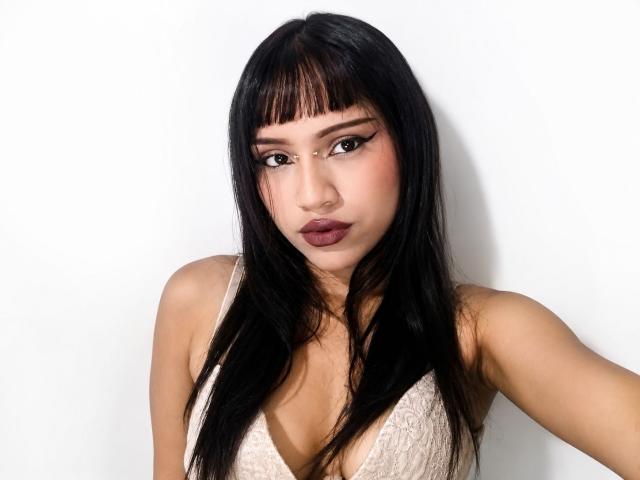 AkariCherise, con lunghi capelli neri, frangia e un'espressione seria, indossa un reggiseno bianco in pizzo, mettendo in mostra il suo seno piccolo su uno sfondo bianco..