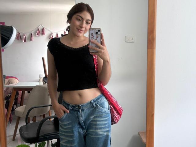 SpencerLe with brown hair, black crop top, showing her flat stomach, and blue jeans, holding a phone with a pink patterned bag on her right shoulder.