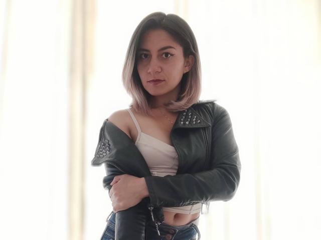 ValeriaLoren, com um corte de cabelo bob em tons de rosa-marrom, vestindo uma jaqueta de couro preta, uma camiseta branca e jeans azuis, braços cruzados, com uma expressão séria, iluminação suave. Seios levemente visíveis sob a camiseta..