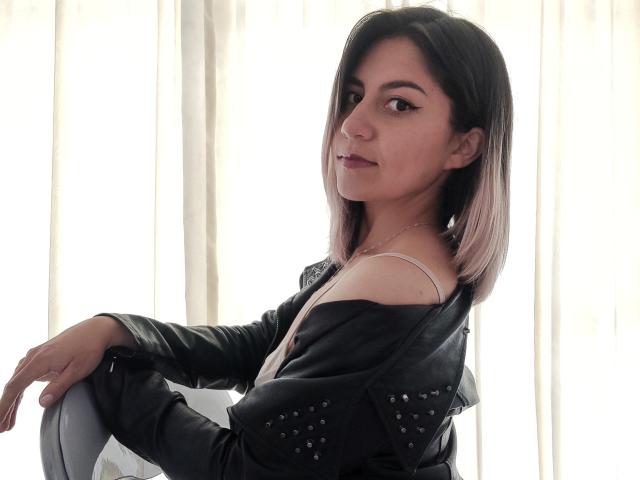 ValeriaLoren mit schulterlangem, schwarz-rosa Haar, trägt eine schwarze Lederjacke und sitzt mit einer Hand auf einem Stuhl, wobei sie über ihre Schulter schaut und ihre kleinen Brüste zeigt..