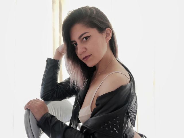 ValeriaLoren, con cabello negro y rosa hasta los hombros, chaqueta de cuero negro, sostén blanco y collar, sentada en una silla blanca, mirando a la cámara con una ligera sonrisa, con la mano en el cabello..