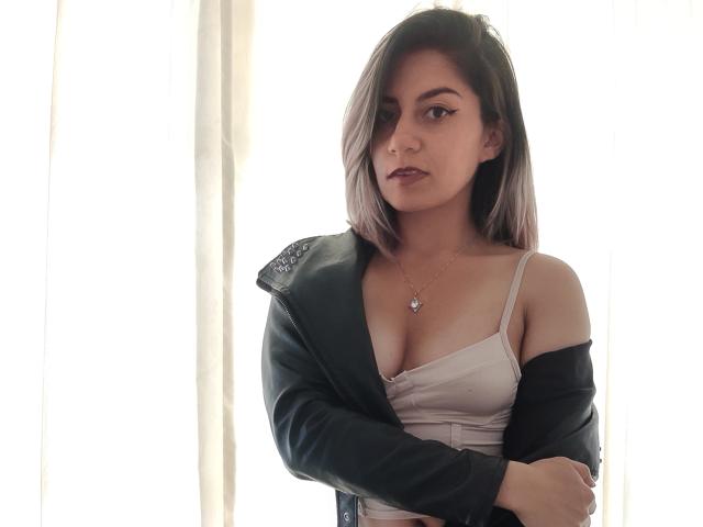 ValeriaLoren, com cabelo castanho na altura dos ombros, jaqueta verde, top branco, mostrando seios pequenos, braços cruzados, expressão séria, colar com um pequeno pingente, em pé sob luz forte..