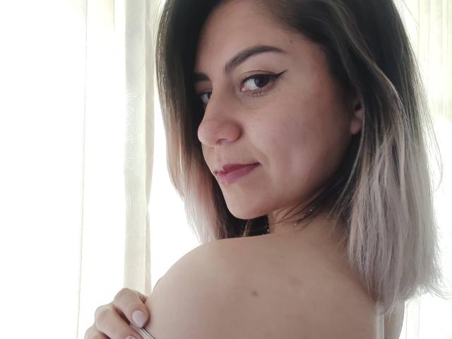 ValeriaLoren, con el hombro visible, ojos marrones claros, delineador negro, labios rosados, cabello ombre marrón oscuro a gris de longitud media, ligera sonrisa, sosteniendo el hombro, luz natural suave..