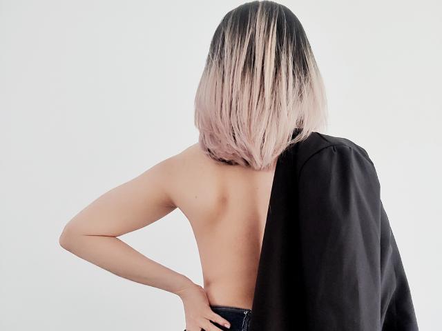 ValeriaLoren, vista da dietro, senza top, con un caschetto biondo-rosa, un giubbotto nero drappeggiato su una spalla, una mano appoggiata sul fianco, pelle chiara, corporatura esile, senza seni o glutei visibili..
