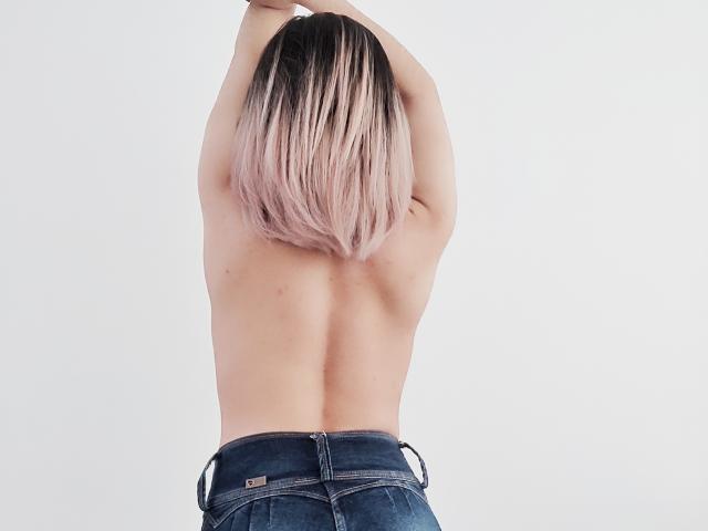 ValeriaLoren, mit erhobenen Armen, oben ohne, mit einem Farbverlauf von rosa zu schwarz in ihrem Haar, trägt blaue Jeans und zeigt ihren nackten Hintern und Rücken, vor einem weißen Hintergrund..