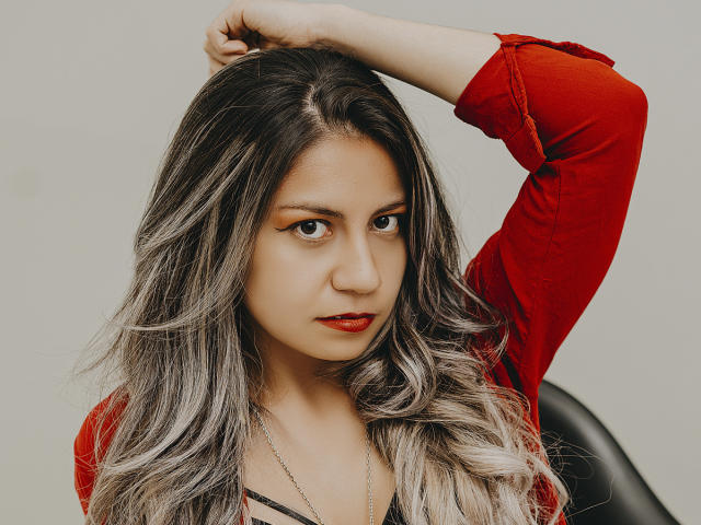 ValeriaLoren, con lunghi capelli grigio-marrone ondulati, labbra rosse e una camicia rossa a maniche lunghe, con un braccio alzato, che guarda la fotocamera, seduta su una sedia nera, con la pelle chiara e un trucco leggero..