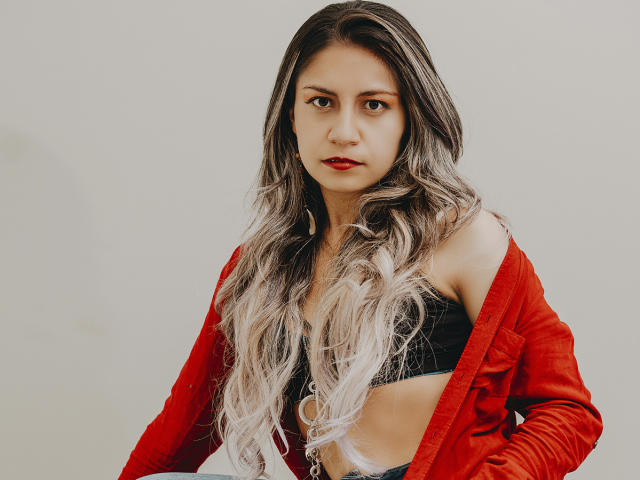 ValeriaLoren mit langem, gewellten, grau-blondem Haar, einem roten Pullover, einem schwarzen Crop-Top und rotem Lippenstift, die direkt in die Kamera schaut und einen ernsten Gesichtsausdruck hat. Ihre Brüste sind sichtbar..