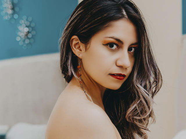 ValeriaLoren hat dunkles, welliges Haar, roten Lippenstift und einen verführerischen Blick. Sie trägt ein rotes Ohrring mit einer goldenen Spirale und ist schulterfrei, wobei ihr Hintern leicht sichtbar ist..